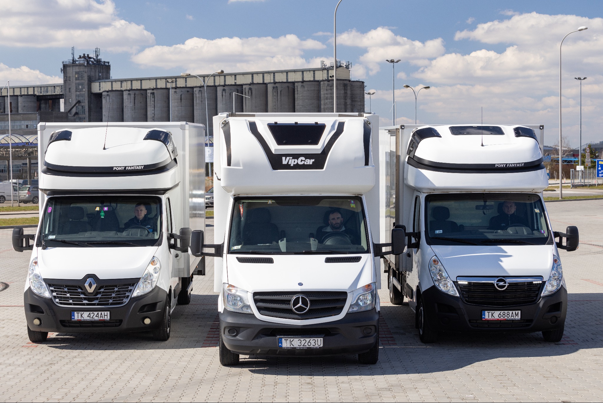 Trzy białe samochody dostawcze różnych marek (Renault, Mercedes, Opel) zaparkowane obok siebie na placu, z widocznymi kierowcami w kabinach, na tle betonowego budynku magazynowego i błękitnego...