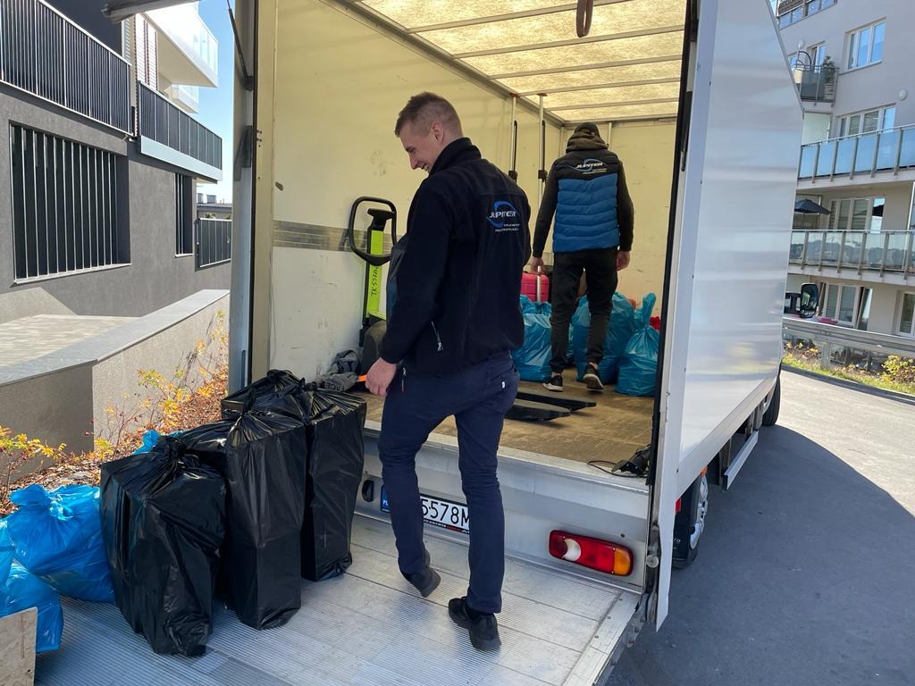 Dwóch pracowników firmy transportowej ładuje worki i kartony do wnętrza samochodu dostawczego na tle nowoczesnego budynku mieszkalnego.