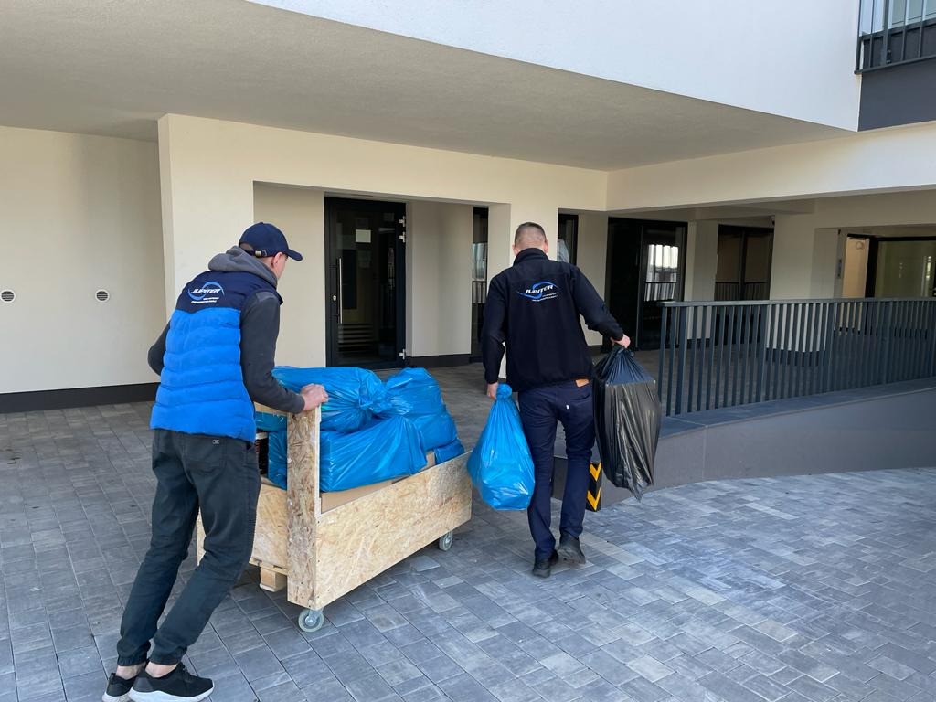 Pracownicy firmy przeprowadzkowej w Kielcach transportują wózkiem pakunki zabezpieczone niebieską folią, jeden z nich niesie dodatkowo czarne worki na śmieci.