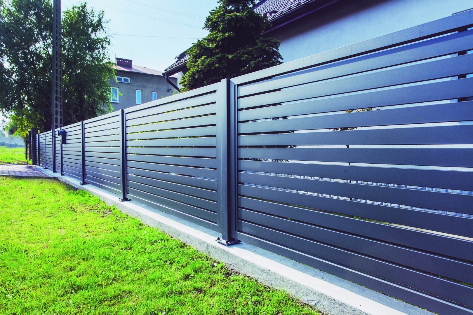 Nowoczesne, poziome przęsła ogrodzenia z metalu w kolorze grafitowym, zamontowane na betonowej podmurówce, otoczone zielonym trawnikiem.