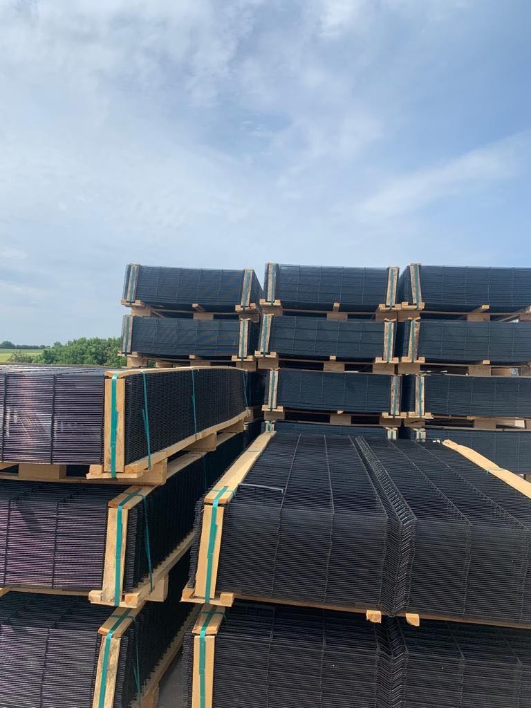 Stosy paneli ogrodzeniowych w kolorze antracytowym, ułożone na drewnianych paletach, zabezpieczone zielonymi taśmami. Tło: błękitne niebo i zieleń drzew.