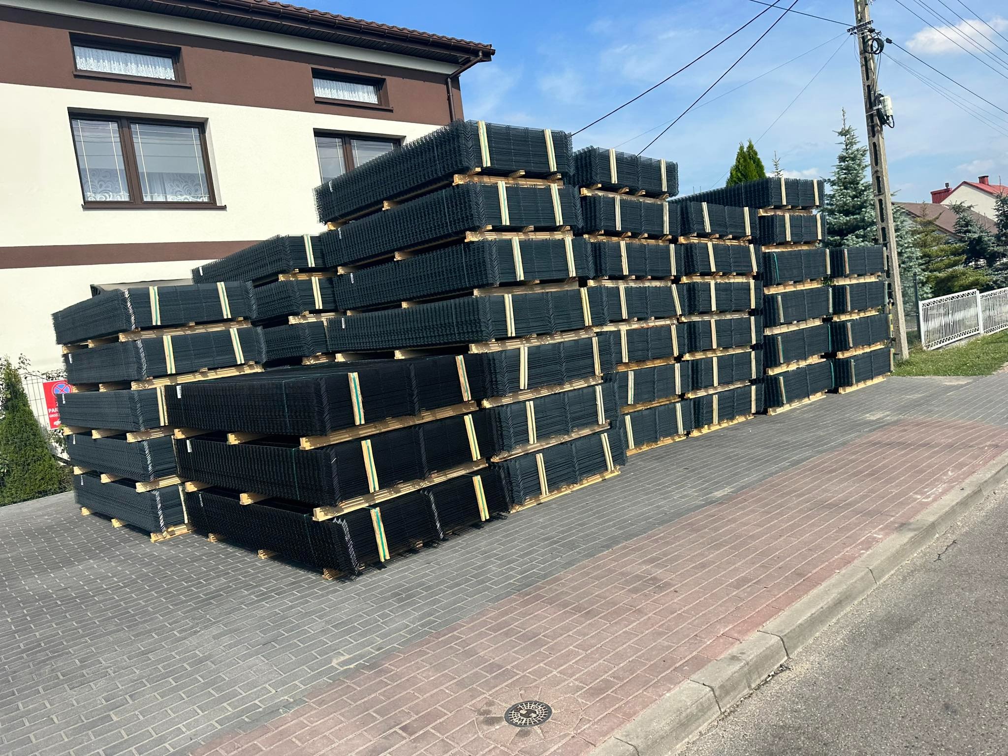Stos paneli ogrodzeniowych na paletach, złożony na chodniku przed budynkiem. Widoczne słupy elektryczne i domy w tle.