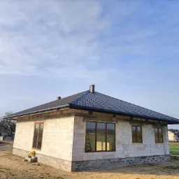 Surowy budynek jednorodzinny z szarym dachem z blachy trapezowej, widoczne obróbki blacharskie, okna z brązowymi ramami, ściany z betonu komórkowego, w tle błękitne niebo z chmurami i budynki...