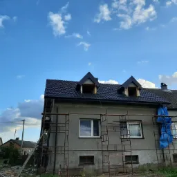 Dom w trakcie wymiany dachu, widoczne rusztowania, nowa, czarna dach&oacute;wka i elementy więźby dachowej w lukarnach, błękitne niebo w tle.