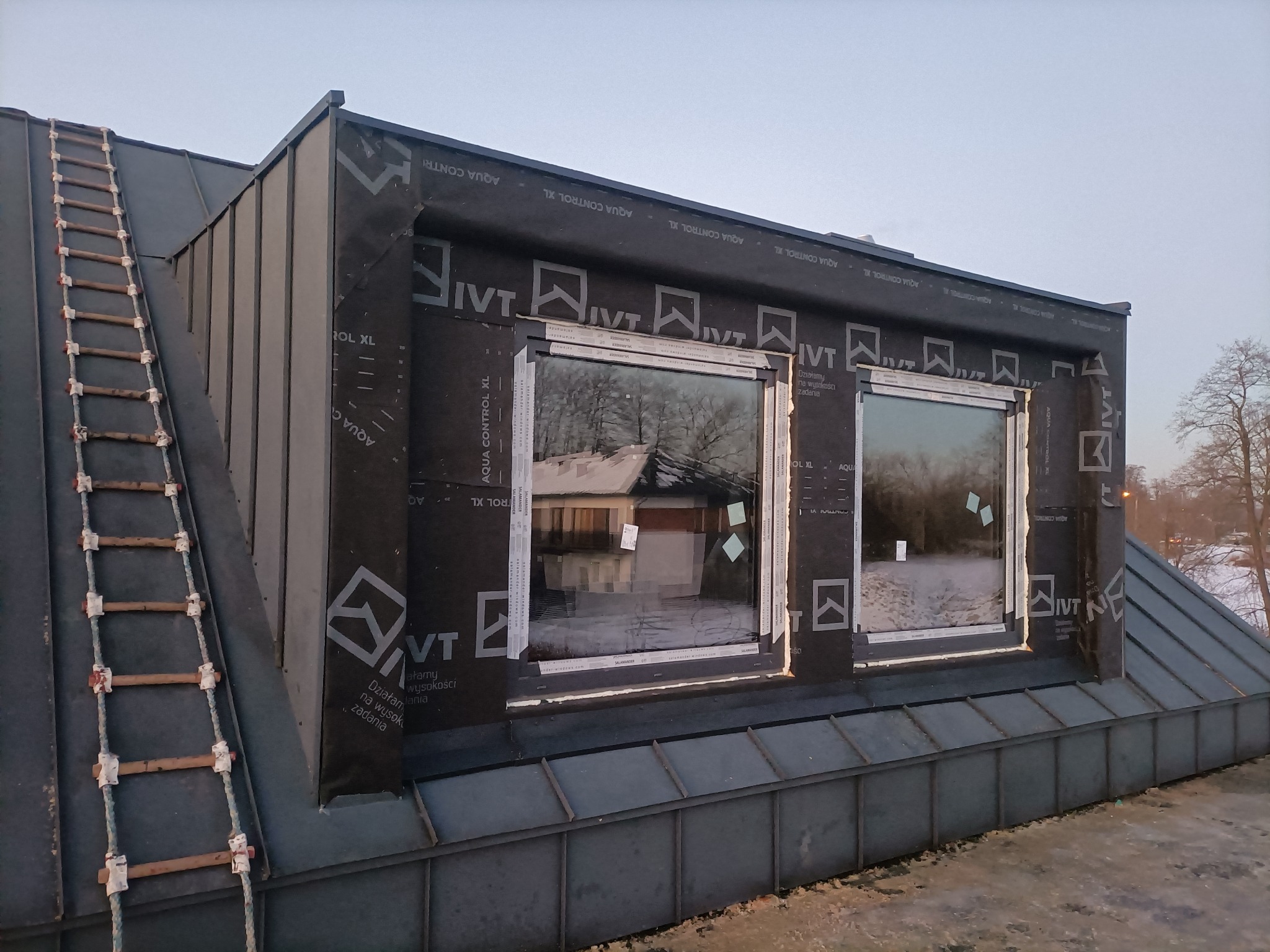 Dach z blachy trapezowej w trakcie montażu okien dachowych, zabezpieczony membraną paroprzepuszczalną z nadrukiem, drabina sznurowa oparta o krawędź dachu.