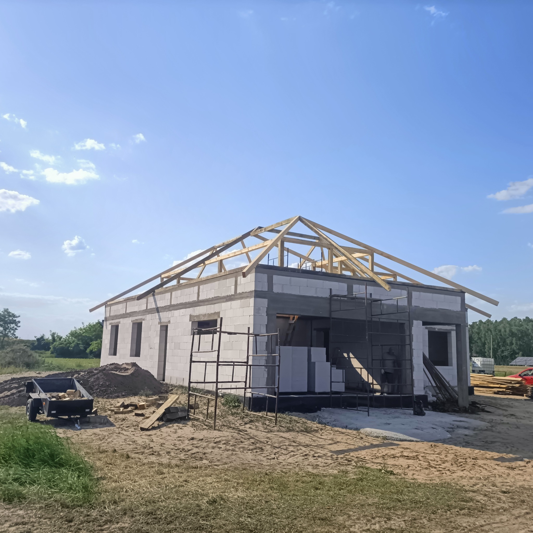 Budowa domu jednorodzinnego w trakcie wznoszenia więźby dachowej, widoczne pustaki ceramiczne, rusztowania i przyczepka z materiałami budowlanymi na placu budowy.