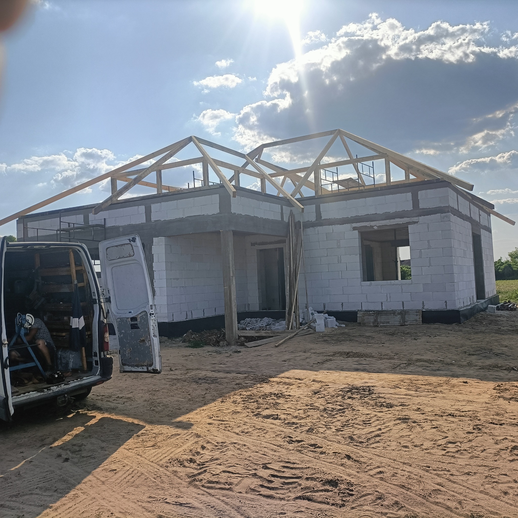 Wznoszony budynek jednorodzinny z białych bloczków, widoczna konstrukcja drewniana dachu, na pierwszym planie częściowo otwarty samochód dostawczy z narzędziami i materiałami budowlanymi, słoneczny...