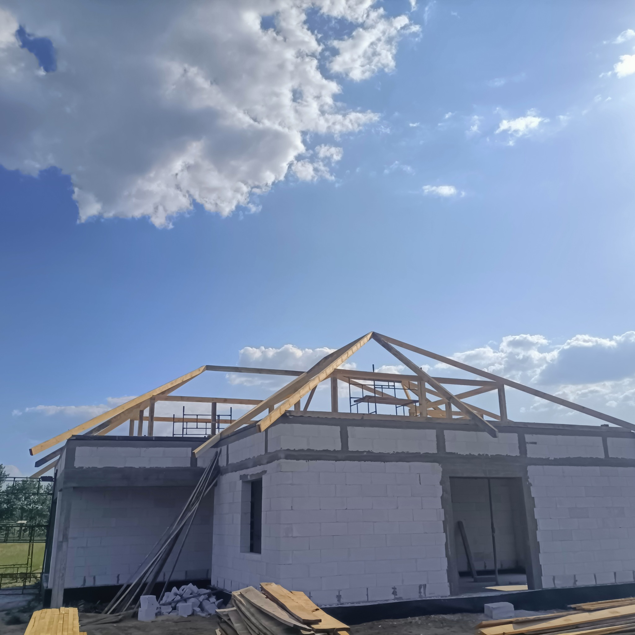 Surowy stan budynku z białych bloczków z widoczną drewnianą konstrukcją więźby dachowej na tle błękitnego nieba z chmurami.