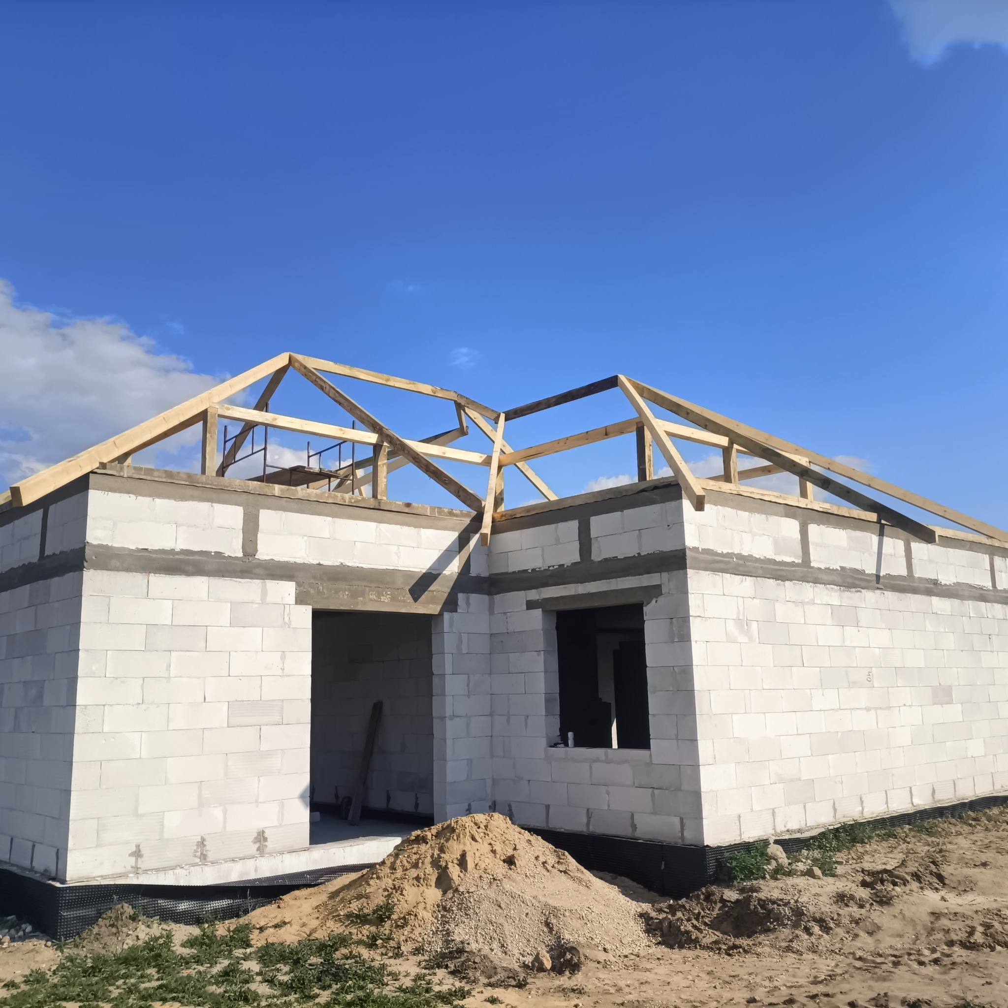 Wznoszenie więźby dachowej z drewnianych belek na budynku z białych bloczków, widoczne rusztowanie i piasek, błękitne niebo w tle.