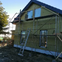 Dom jednorodzinny w trakcie prac związanych z izolacją ścian zewnętrznych, widoczne rusztowania, okna zabezpieczone folią, oraz fragment nowo położonego, ciemnego dachu.