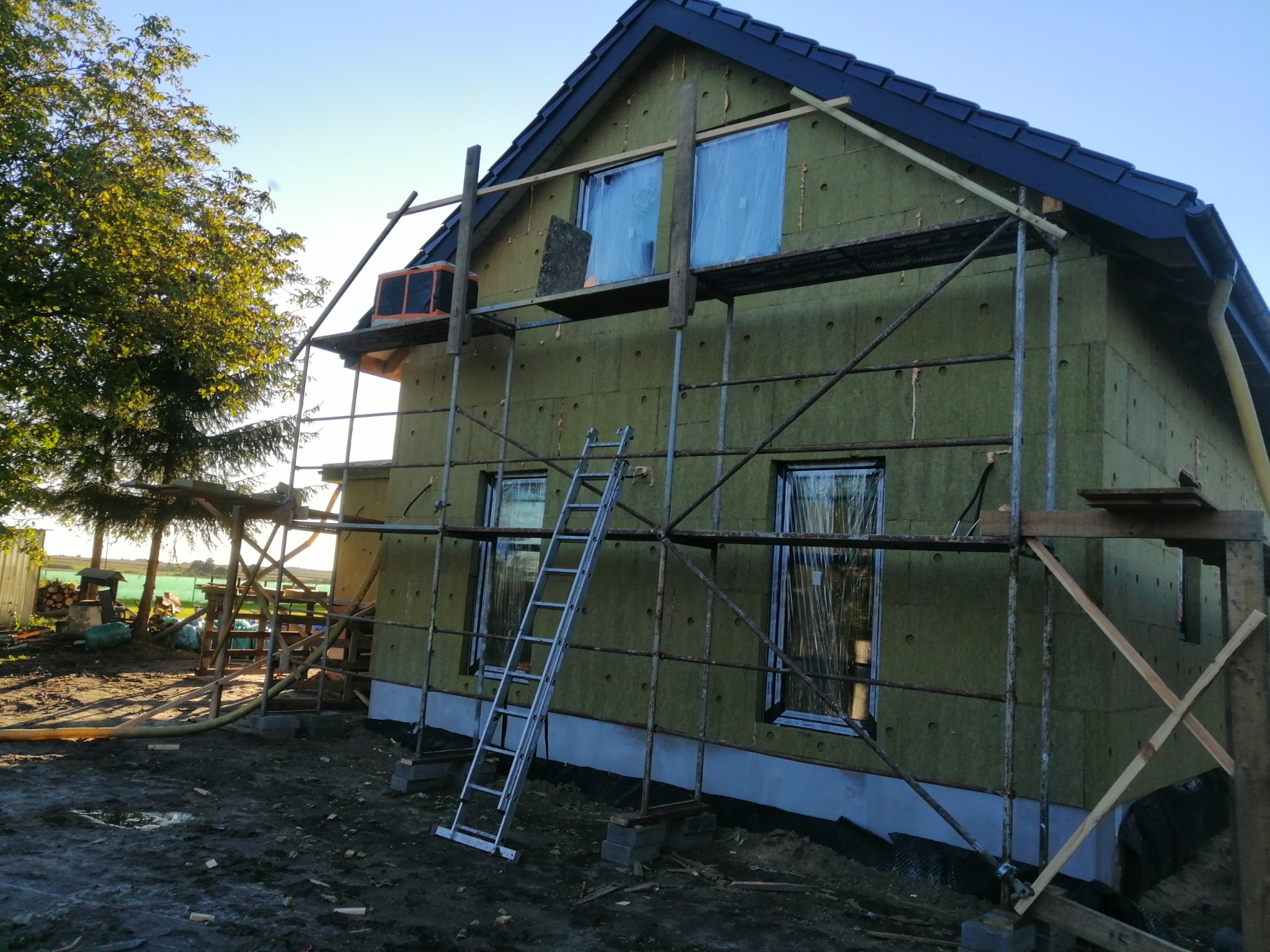 Dom jednorodzinny w trakcie prac związanych z izolacją ścian zewnętrznych, widoczne rusztowania, okna zabezpieczone folią, oraz fragment nowo położonego, ciemnego dachu.