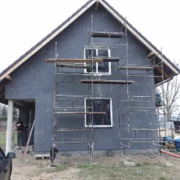 Dom jednorodzinny w trakcie ocieplania, pokryty szarą warstwą izolacyjną, z widocznym rusztowaniem i oknami zabezpieczonymi folią, pracownicy w trakcie wykonywania prac.