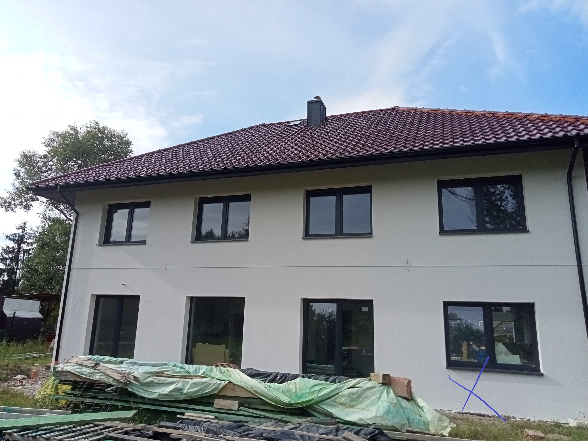 Dwukondygnacyjny dom w trakcie budowy z widocznym rusztowaniem i materiałami budowlanymi na pierwszym planie, tynkowana elewacja i ciemna stolarka okienna.