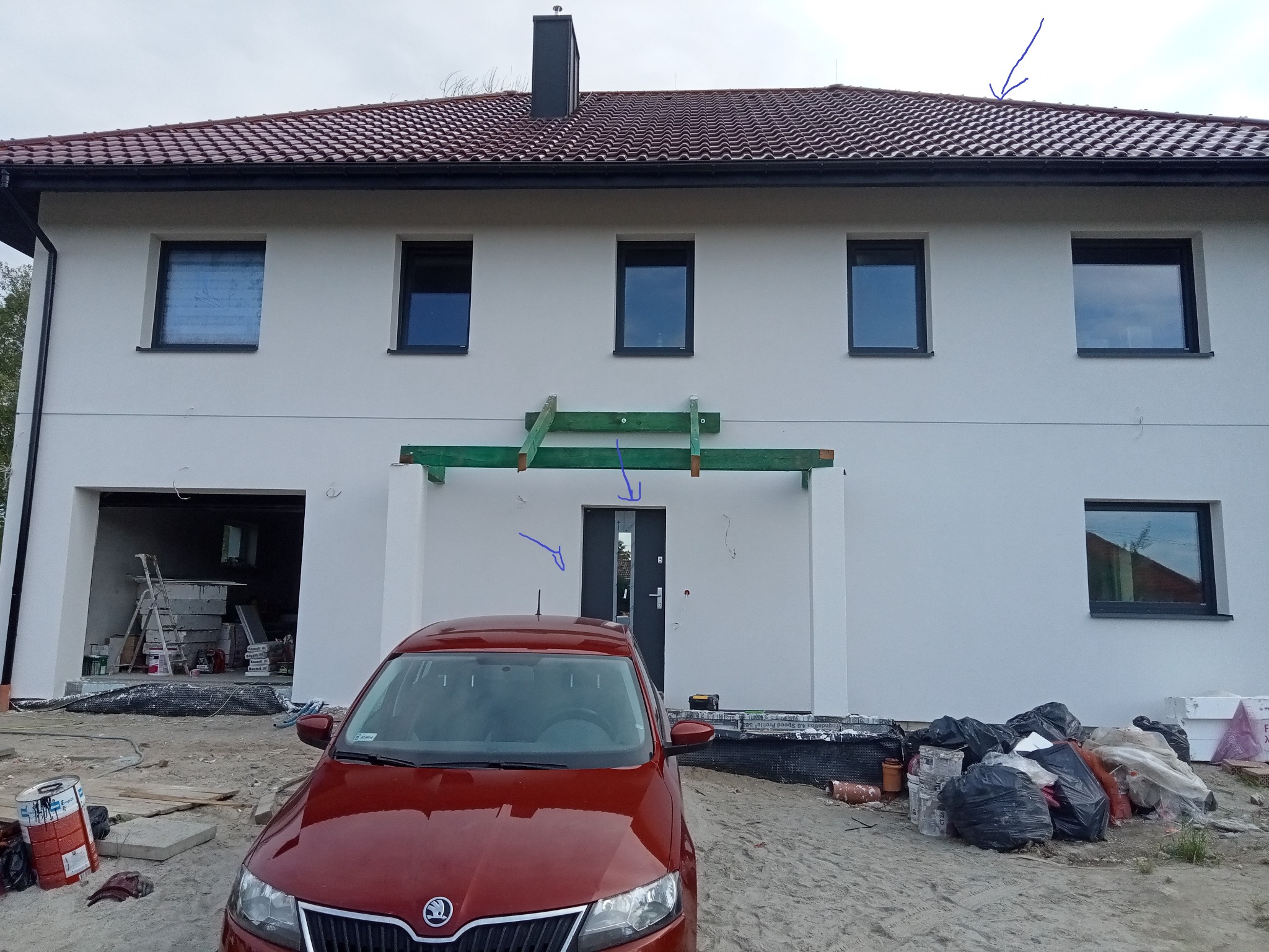 Dwupiętrowy dom w trakcie budowy z czerwonym samochodem na podjeździe, widoczne rusztowania i materiały budowlane, elewacja biała, dach z czerwonej dachówki.