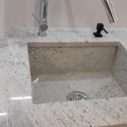 Jasny blat kuchenny z wbudowanym kwadratowym zlewem, bateria i dozownik na płyn do naczyń, widoczny odpływ.