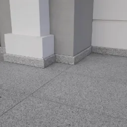 Szara, granitowa posadzka na zewnątrz budynku, z obrzeżem z tego samego materiału wokół filarów i ściany. Widoczny fragment czarnych drzwi po lewej stronie.