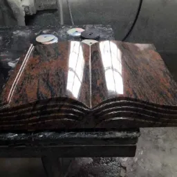 Otwarta księga wykonana z polerowanego, brązowo-czarnego granitu, leżąca na warsztatowym stole obok tarcz szlifierskich.