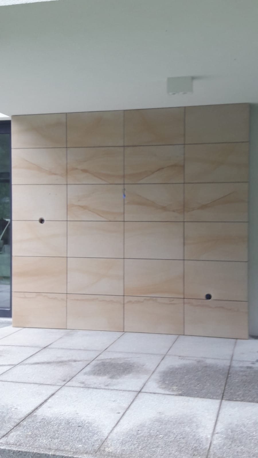 Ściana zewnętrzna wyłożona jasnymi płytami imitującymi piaskowiec, z widocznymi otworami na instalacje elektryczne, przylegająca do szarego tarasu.