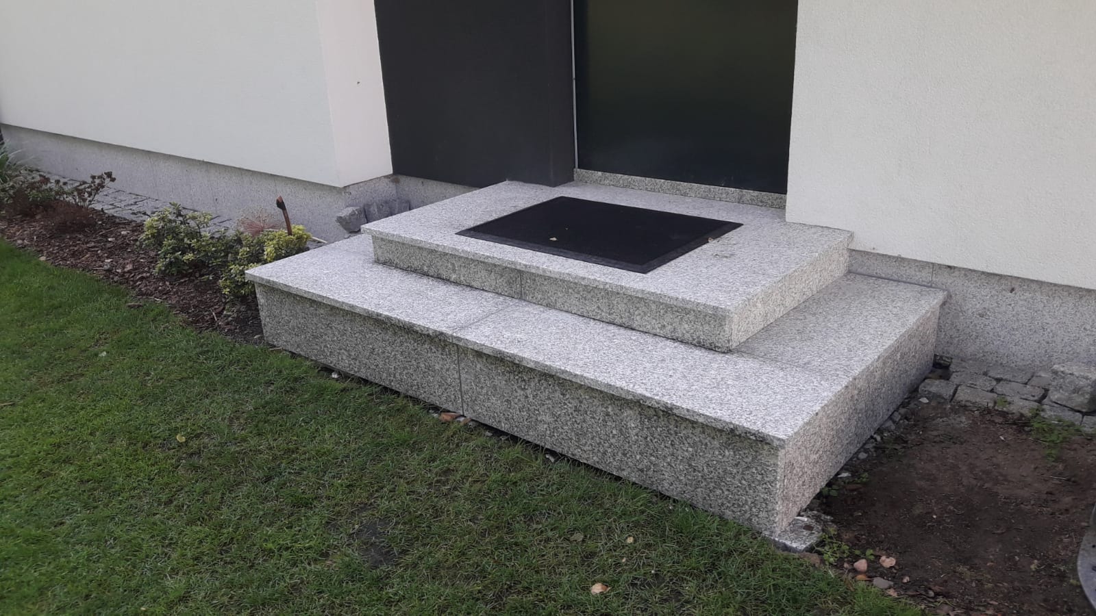 Dwu-stopniowe wejście do domu wyłożone jasnoszarym kamieniem z widoczną czarną wycieraczką na górnym stopniu, otoczone trawnikiem i roślinnością.