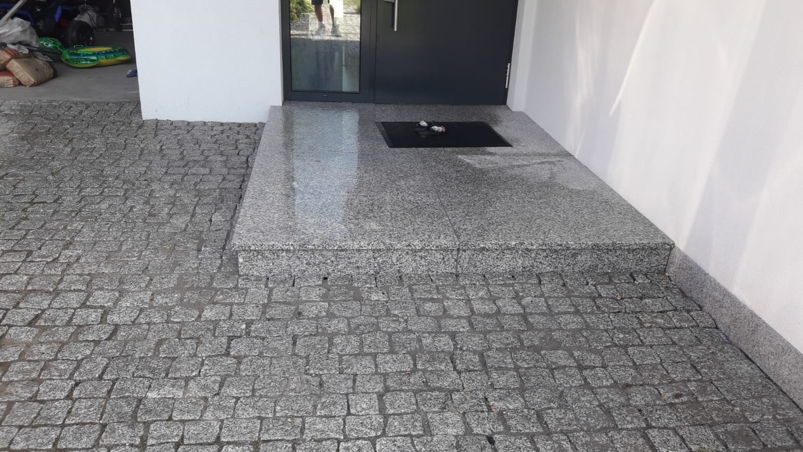 Granitowe schody wejściowe z kostki brukowej i płyt z czarną wycieraczką przed drzwiami.