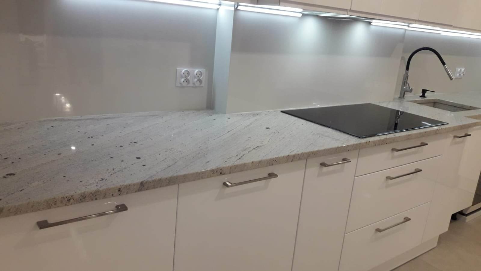 Jasny blat z kamienia naturalnego o delikatnym wzorze, wkomponowany w nowoczesną białą zabudowę kuchenną z płytą indukcyjną i chromowanymi uchwytami.