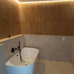 Nowoczesna łazienka z drewnianą boazerią, podświetleniem LED, białą wanną wypełnioną wodą i czarną armaturą.