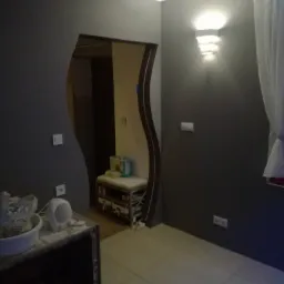 Nowoczesne wnętrze z szarą ścianą, ozdobnym oświetleniem ściennym i wejściem o nieregularnym kształcie. Widoczne gniazdka elektryczne, biała zasłona i fragment podłogi wyłożonej płytkami.