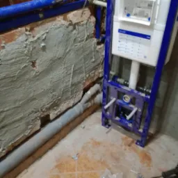 Stelaż podtynkowy WC Geberit Duofix zamontowany przy nierównej, częściowo otynkowanej ścianie z widocznymi rurami kanalizacyjnymi i instalacją wodną, na brudnej podłodze w trakcie prac instalacyjnych.