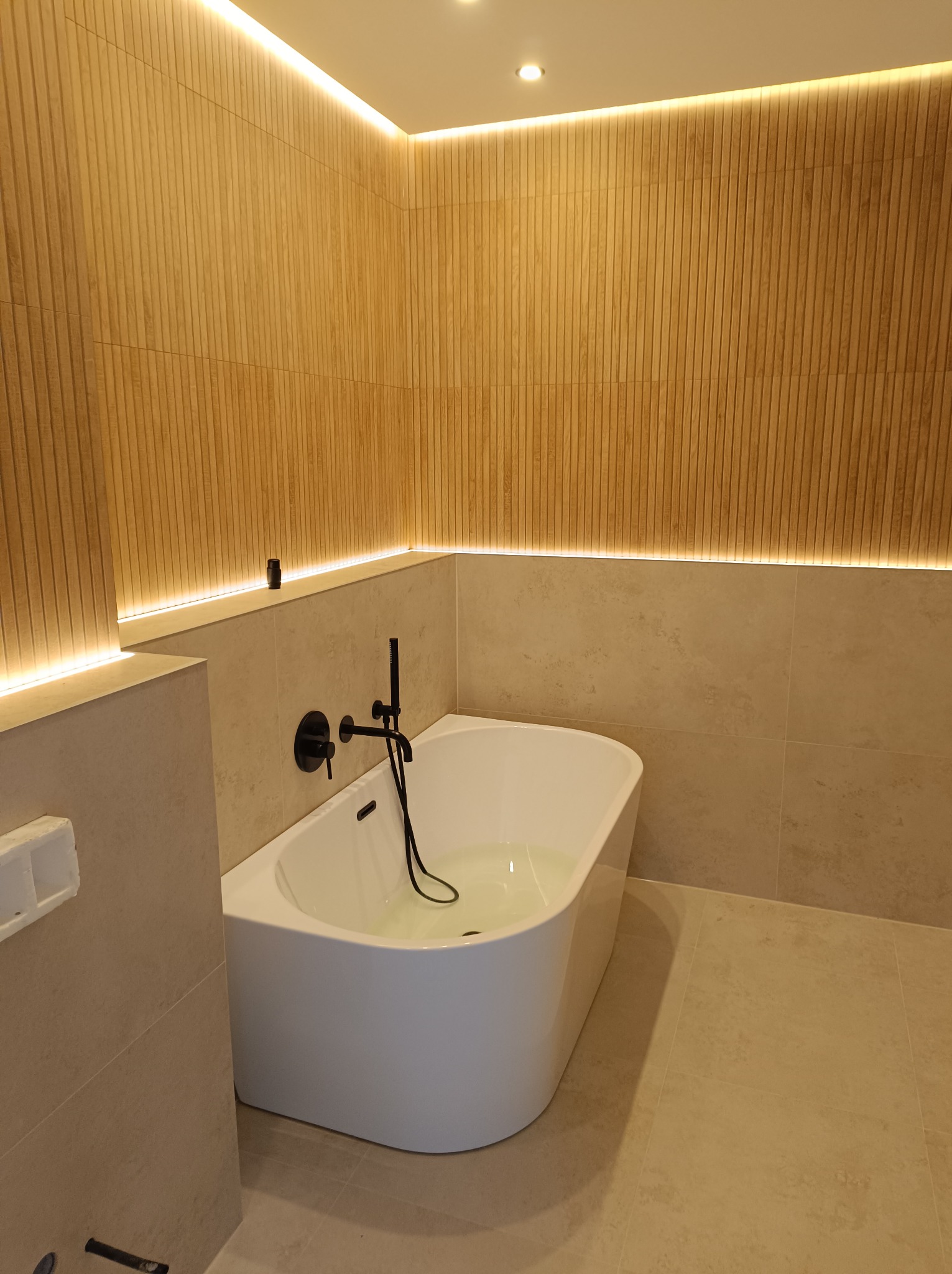 Nowoczesna łazienka z wolnostojącą wanną wypełnioną wodą, czarną armaturą i podświetlanymi panelami ściennymi imitującymi drewno.