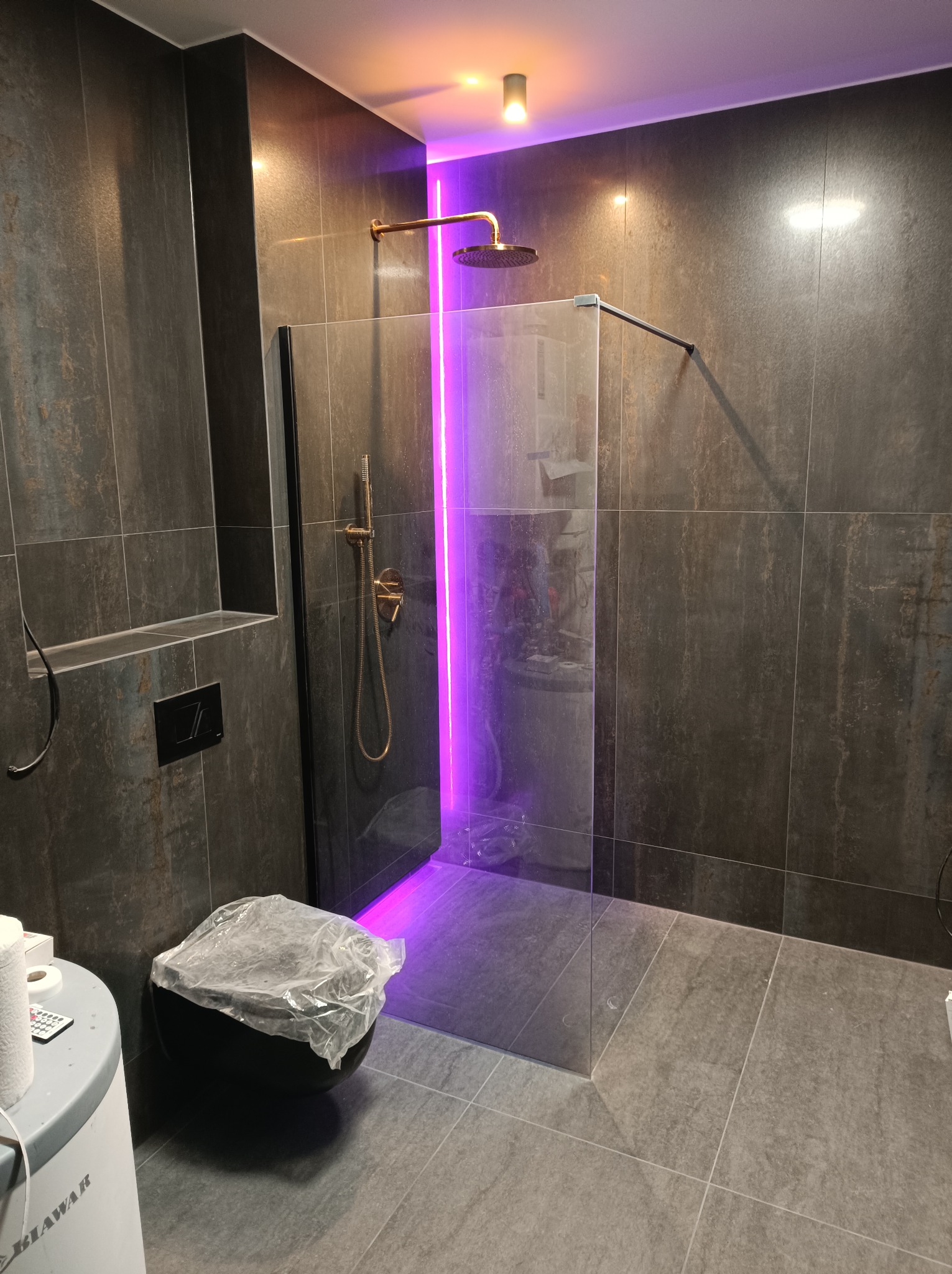 Nowoczesna łazienka z kabiną prysznicową typu walk-in, ciemna okładzina ścienna, deszczownica w kolorze złotym, podświetlenie LED w kolorze fioletowym, zabezpieczona folią toaleta w trakcie prac.