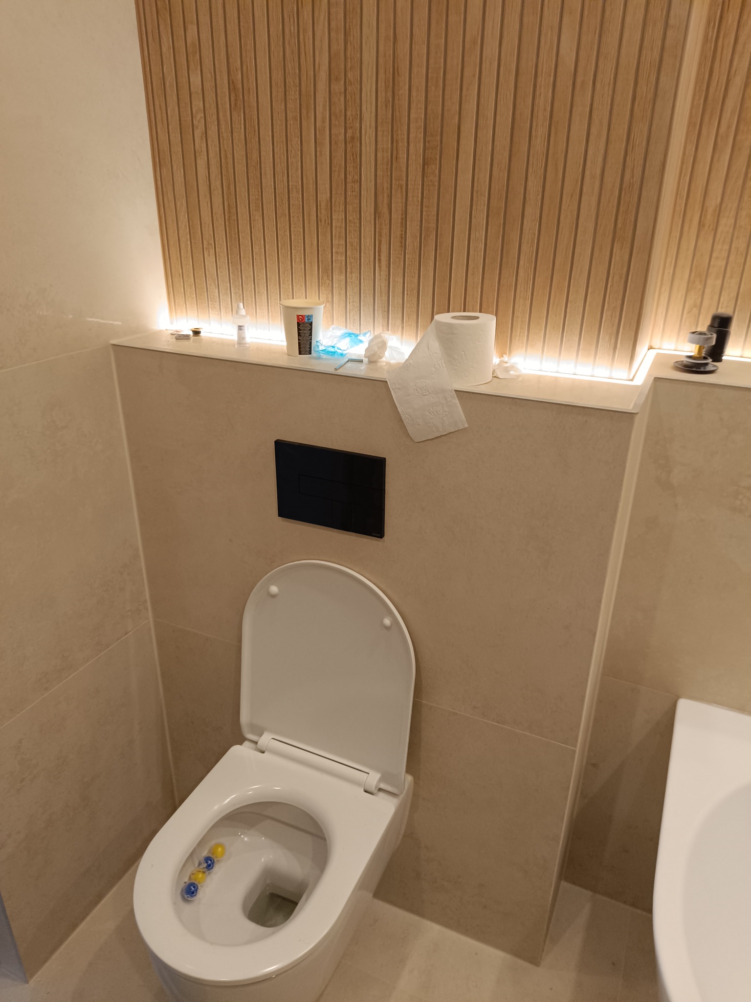 Nowoczesna toaleta z deską podniesioną, z widocznymi kostkami do WC, kremowe płytki na ścianach, drewniana okładzina z podświetleniem LED w górnej części ściany, akcesoria łazienkowe na półce.