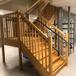 Jasne, drewniane schody z czarną, metalową poręczą w przestrzeni publicznej, widoczne filary i oznaczenia kierunkowe.