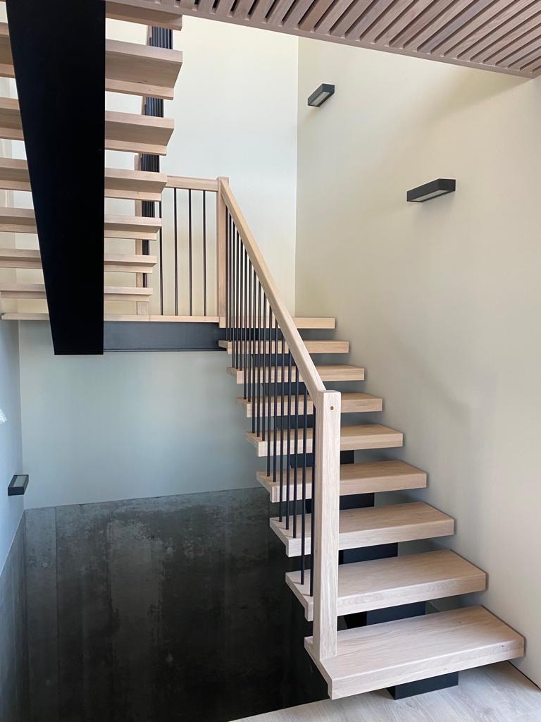 Nowoczesne schody z jasnego drewna z czarnymi metalowymi balustradami i podstopnicami, oświetlenie punktowe na ścianie, widok na betonową piwnicę.