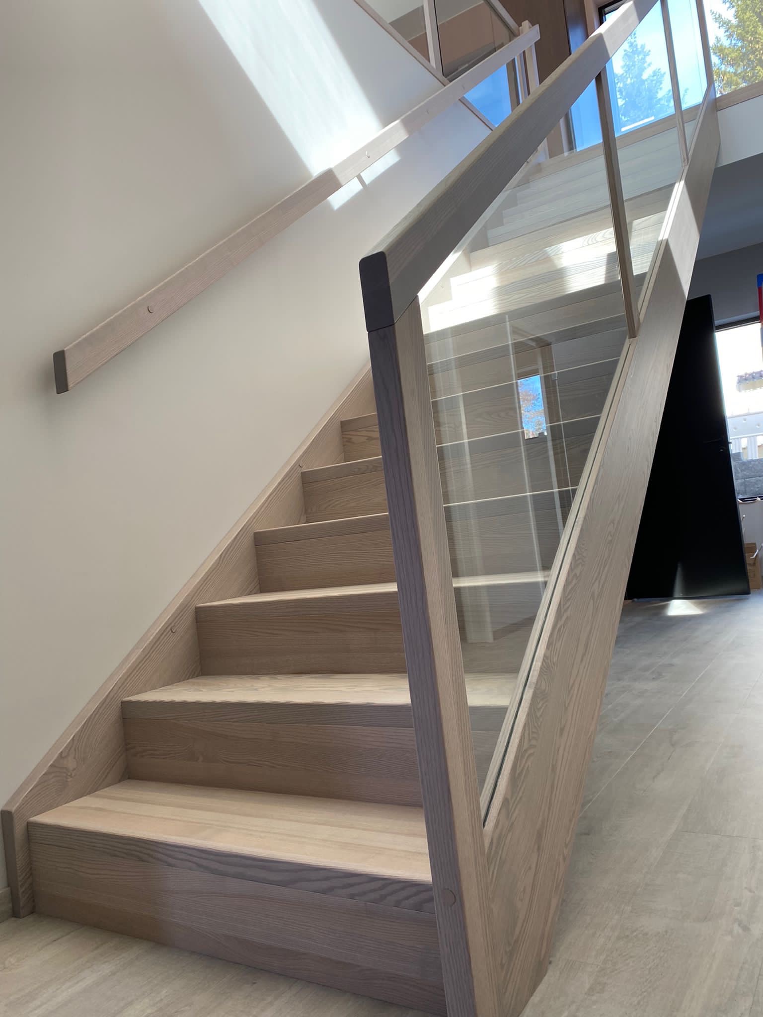 Nowoczesne schody z jasnego drewna z przeszkloną balustradą, widok z dołu, światło słoneczne odbija się od ściany.