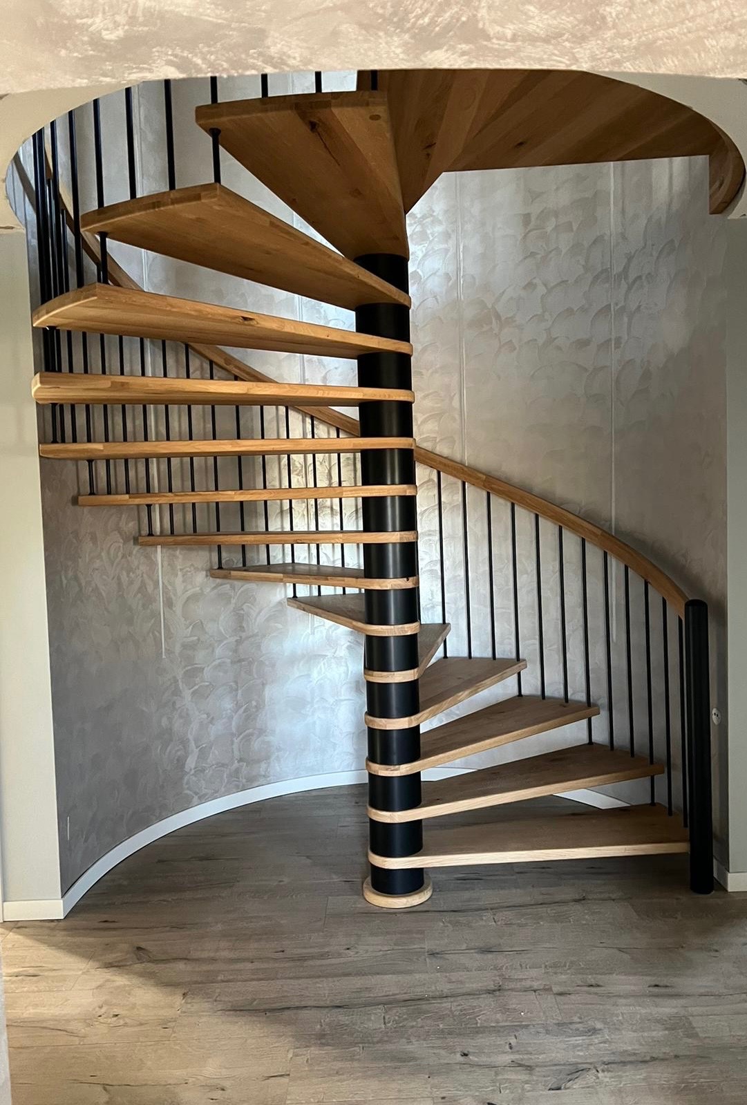 Kompaktowe, spiralne schody z drewnianymi stopniami i czarną metalową konstrukcją na tle ściany z dekoracyjną fakturą.