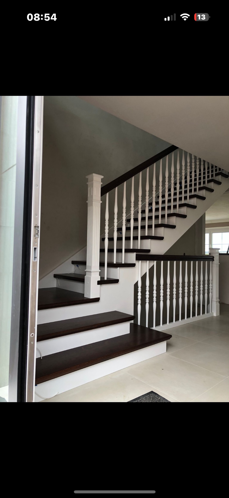 Nowoczesne schody z ciemnego drewna i białymi balustradami, widoczne z dołu z otwartymi drzwiami po lewej stronie, oświetlone naturalnym światłem.