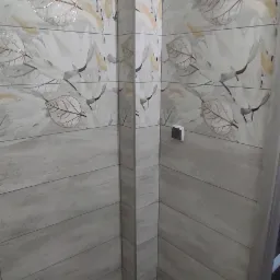 Wykończona ściana i podłoga w łazience z szarymi płytkami imitującymi drewno i dekoracyjnym motywem liści, widoczny narożnik zabudowany płytkami, gniazdko elektryczne na ścianie.