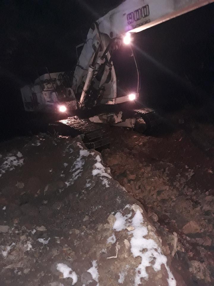 Biała koparka gąsienicowa z włączonymi światłami pracuje w nocy, widoczna ziemia z resztkami śniegu.