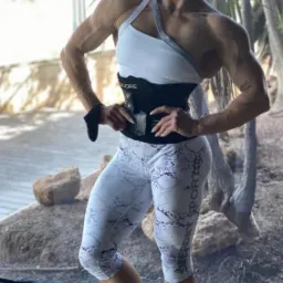 Kobieta w sportowym stroju, prezentująca wyrzeźbioną sylwetkę, pozuje w pomieszczeniu z widokiem na egzotyczną roślinność, ubrana w biało-szary top, wzorzyste legginsy i czarny pas wyszczuplający.