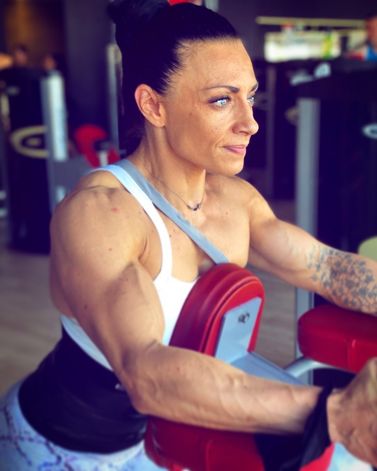Kobieta z mocno rozwiniętymi mięśniami ramion ćwiczy na maszynie na siłowni, widoczny tatuaż na ramieniu i żyły.