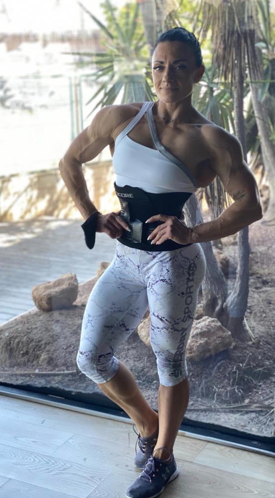 Kobieta w sportowym stroju, prezentująca wyrzeźbioną sylwetkę, pozuje w pomieszczeniu z widokiem na egzotyczną roślinność, ubrana w biało-szary top, wzorzyste legginsy i czarny pas wyszczuplający.