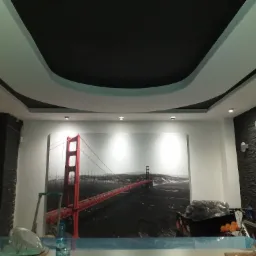 Czarno-biały sufit podwieszany z wbudowanym oświetleniem i dekoracyjną wnęką w kształcie łuku, na ścianie fototapeta z mostem Golden Gate, widoczne elementy wyposażenia i narzędzia podczas prac...