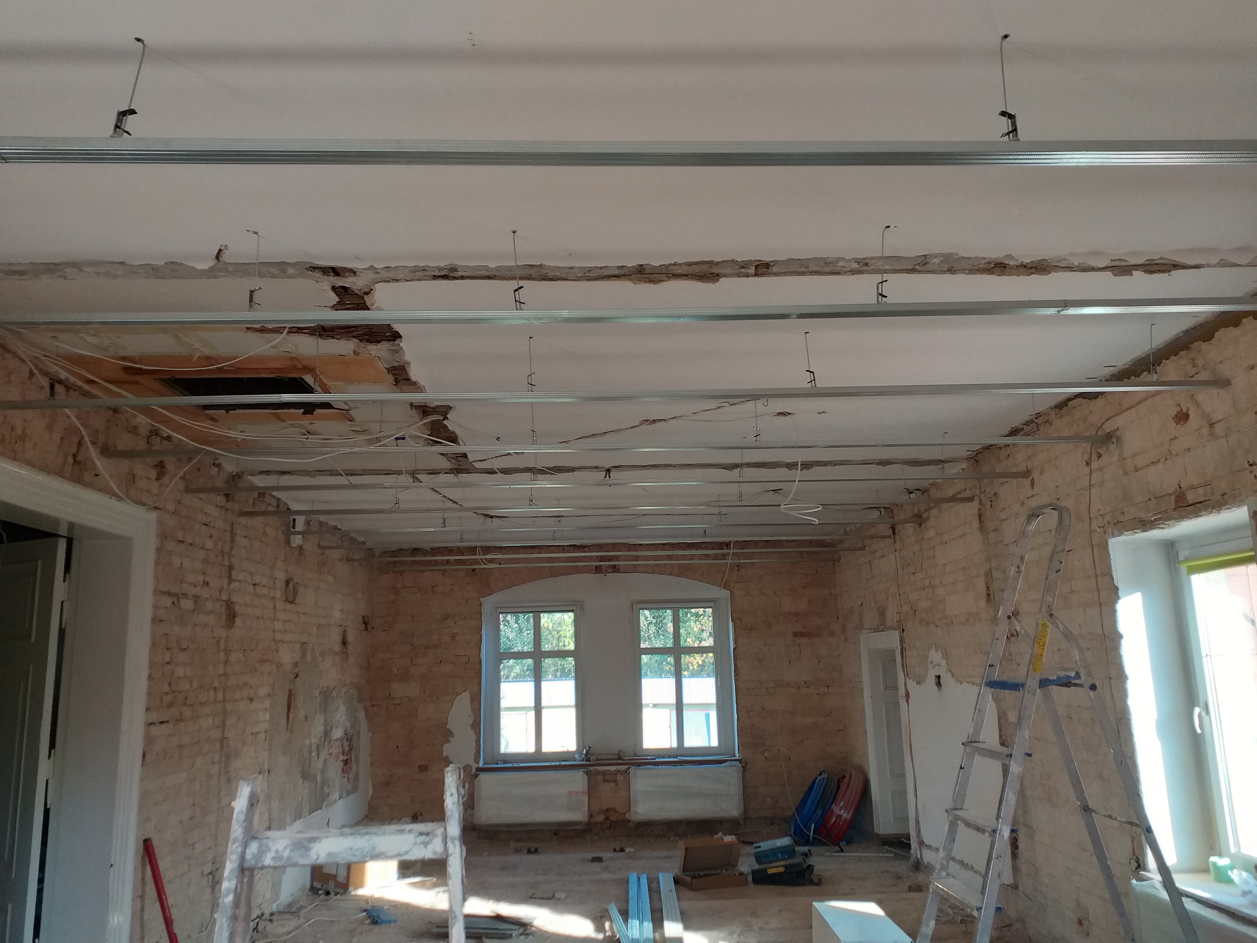 Montaż profili sufitowych w pomieszczeniu z odsłoniętą cegłą, widoczne kable i fragmenty starego sufitu, drabina oparta o ścianę.