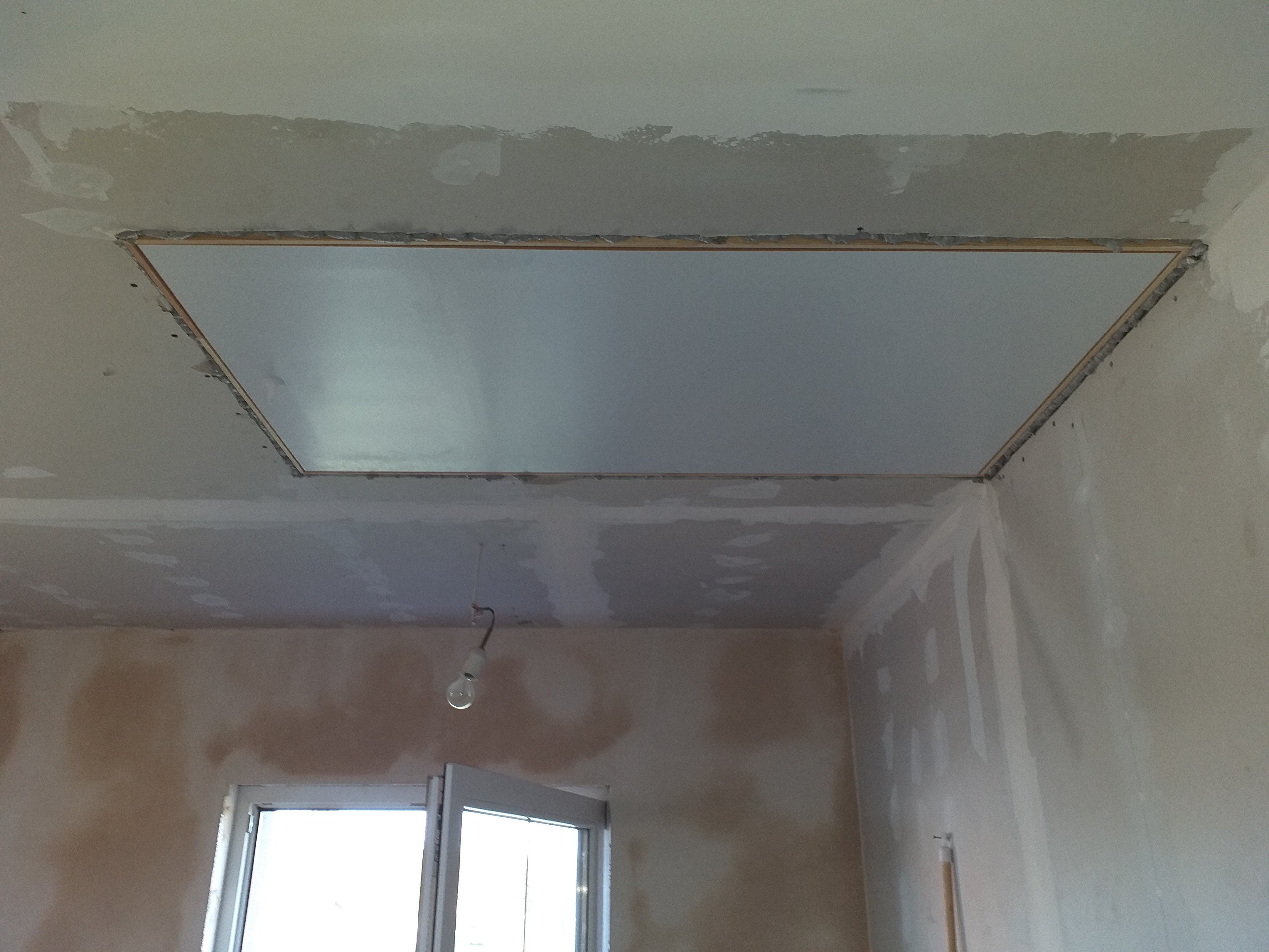 Niedokończony sufit z widoczną konstrukcją nośną, szpachlowanymi ścianami i wiszącą żarówką.