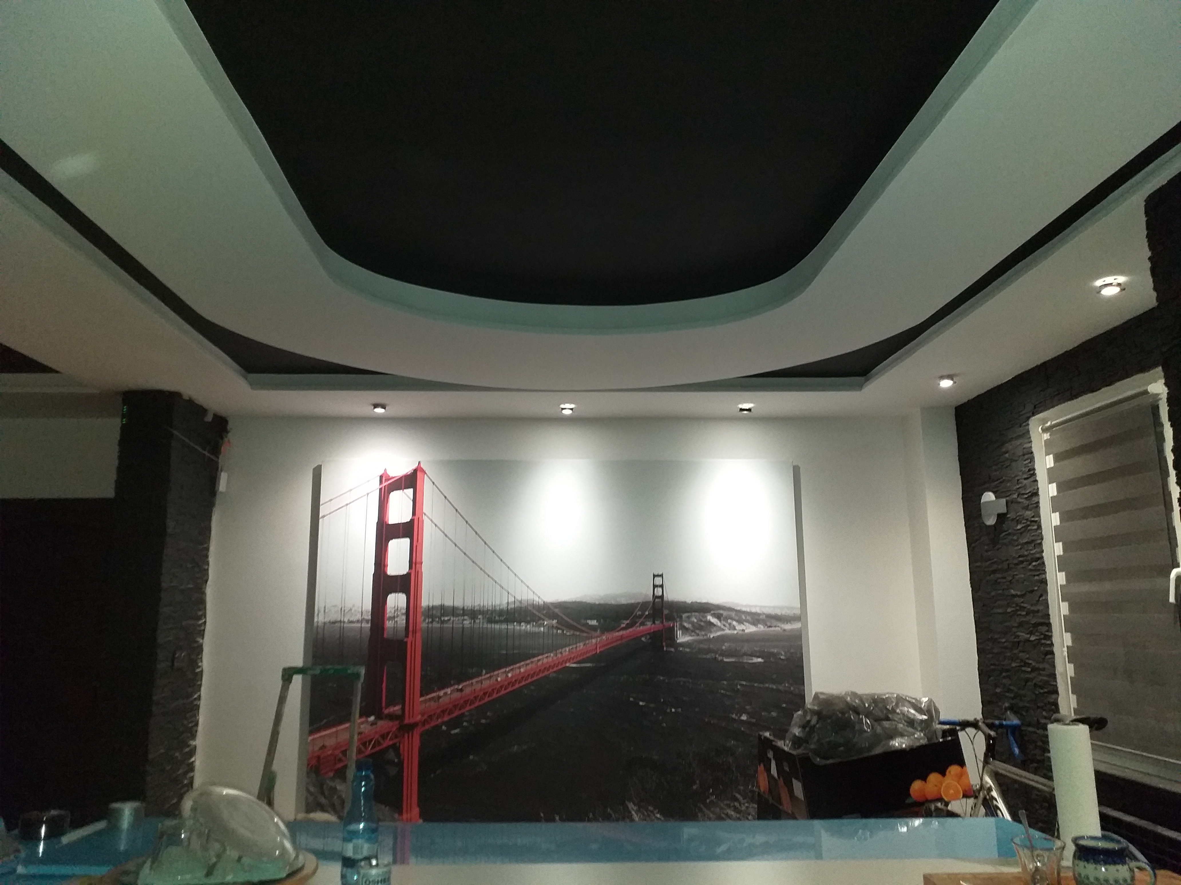 Czarno-biały sufit podwieszany z wbudowanym oświetleniem i dekoracyjną wnęką w kształcie łuku, na ścianie fototapeta z mostem Golden Gate, widoczne elementy wyposażenia i narzędzia podczas prac...