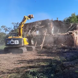 Żółta koparka JCB podczas demontażu ceglanego budynku, w tle błękitne niebo i drzewa, widoczne fragmenty dachu i ścian w trakcie rozbiórki.