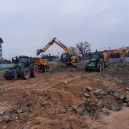 Dwa traktory z przyczepą i dwie koparki JCB na placu budowy z gruzem i ziemią, przygotowujące teren pod inwestycję.