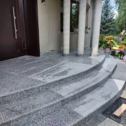 Zewnętrzne schody wejściowe do domu, wyłożone szarym kamieniem z widoczną strukturą ziarnistą, prowadzące do ciemnych drzwi wejściowych z długim, pionowym uchwytem.