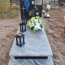 Czarny granitowy pomnik z niebieskim akcentem w kształcie rombu, zdjęciami i kwiatami, ozdobiony dwoma lampionami, na cmentarzu z ławkami w tle.