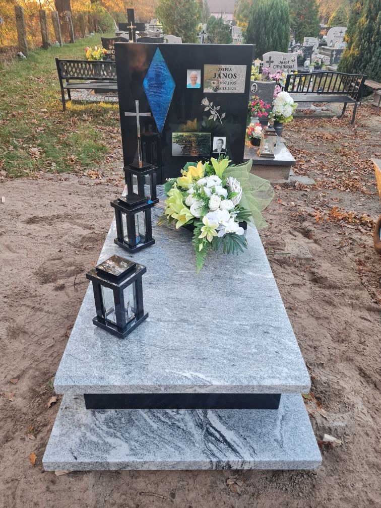 Czarny granitowy pomnik z niebieskim akcentem w kształcie rombu, zdjęciami i kwiatami, ozdobiony dwoma lampionami, na cmentarzu z ławkami w tle.
